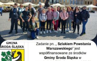 zdjęcie grupowe przy pomniku syrenki na rynku starego miasta w Warszawie, pod spodem herb gminy Środa Śląska i napis "Zadanie pn. >>Szlakiem Powstania Warszawskiego